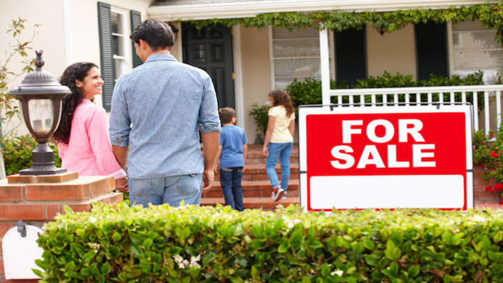 A family walking into a house with a for sale sign in the front yard.