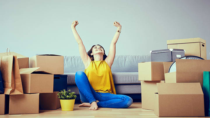 A relieved woman sitting in front of a couch surrounded by moving boxes.