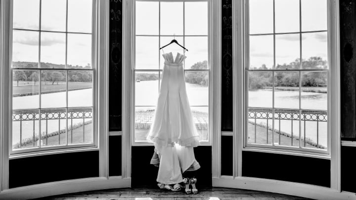 A wedding dress hanging in front of three large bay windows.