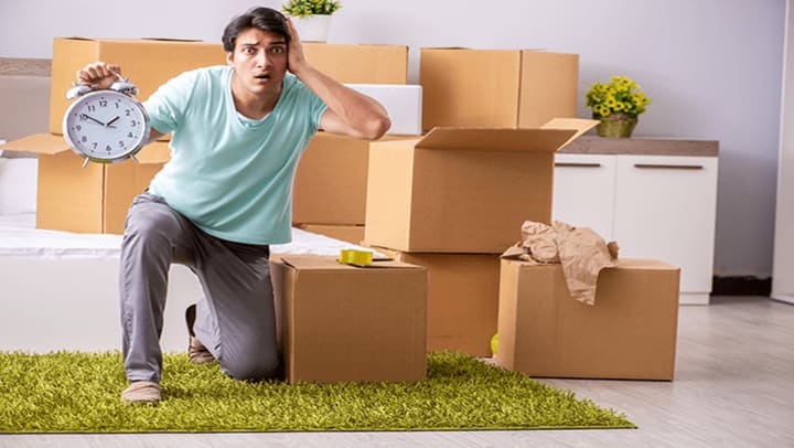 A confused man holding a clock surrounded by moving boxes.
