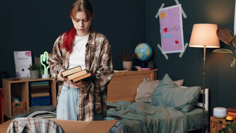 Teenage girl stands in dorm bedroom holding books, packing belongings into boxes, getting ready to move out