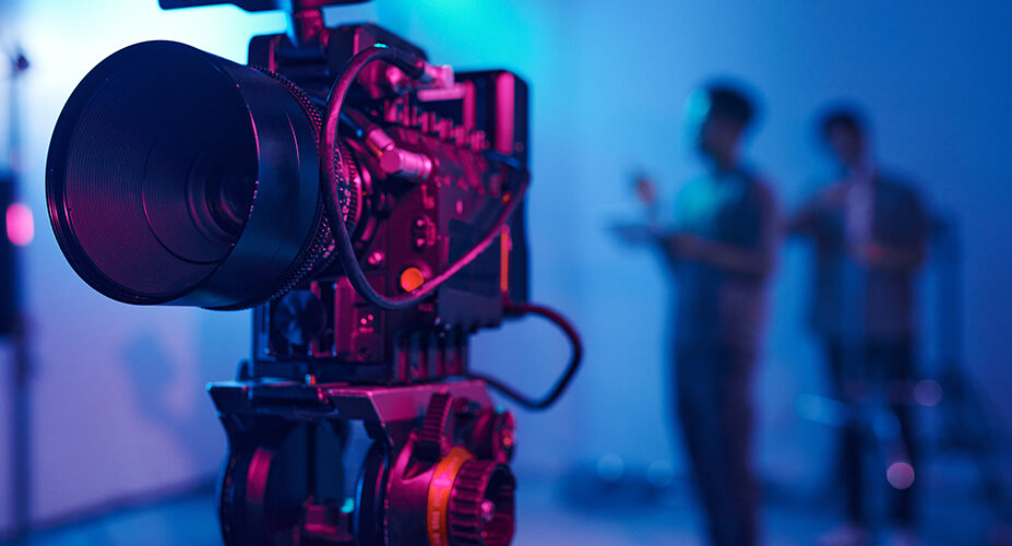 Large camera in focus on movie set or studio with film production crew in the background directing